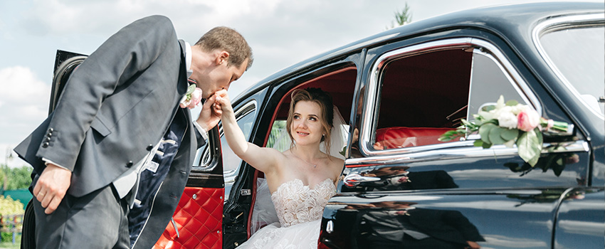 A groom helps a smiling bride out of a vintage car, kissing her hand.